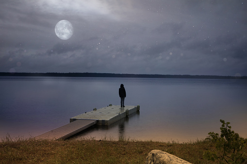 Woman standing alone on the dock of a 
 lake at night under the moon light