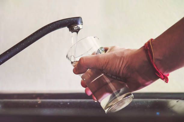 jovem, enchendo um copo com água na pia da cozinha em casa, uma homem derrama água da torneira em um copo de vidro transparente para beber. - pouring water carafe glass - fotografias e filmes do acervo