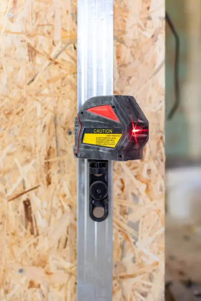 Vertical close up photo of switched-on laser level tool with a red beam stand against room with oriented strand board panel
