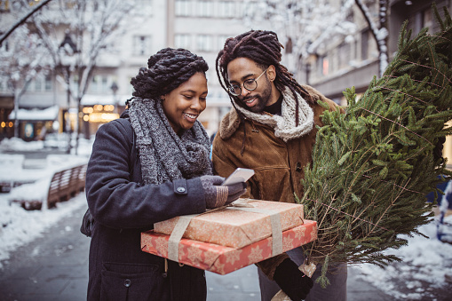 Young couple on Christmas market. They walking around and picking what to buy. They use mobile phone to find best retail. Holding gift boxes and Christmas tree