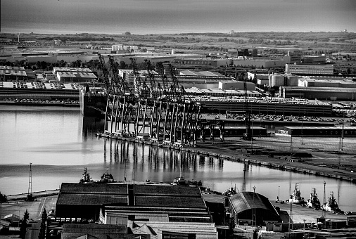 Photo taken from the heights of the city of Barcelona (Spain)\nView of the commercial port.\nUnloading cranes, commercial and fishing boats, car parks, buildings on the Mediterranean port. \nAll the elements are reflected in the water of the port. In the background, the Barcelona countryside.Black and white image
