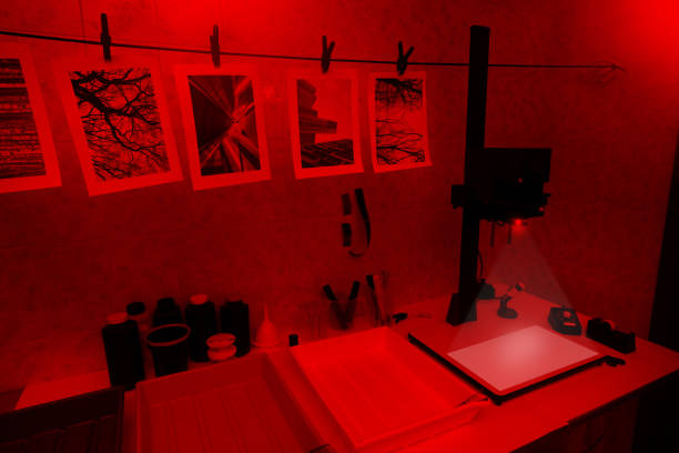 Darkroom for analog photography View of a darkroom with printing tools and materials for analog photography, illuminated by a red light. Photo enlarger projecting light on its base. Also visible are an easel, focusing magnifier, enlarger timer and negative carrier. Tanks filled with chemicals and various tools fill the table. Bottles with developer bath, stop bath and fixer. Black and white photographs hanging on a wire, waiting for complete drying. Some piece of negative film are visible on the background. All pictures on paper constitute personal work. darkroom photos stock pictures, royalty-free photos & images