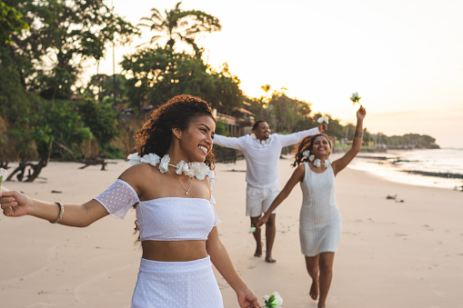 Happy friends celebrating reveillon on the beach, running and holding white flowers. They wear white clothes. Group of young people enjoying and partying together. Happiness, togetherness, youth and new year's eve concepts. Paraiso beach, Mosqueiro