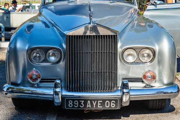 A detailed shot of the iconic Rolls-Royce hood figure perched above a gleaming chrome grille. The image conveys luxury, heritage, and timeless automotive elegance through its polished metal surfaces and clean blue sky backdrop.