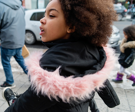 Real mixed-race family with young children shopping in holiday season. They are dressed in winter clothes, and are walking on a city street. Portrait of beautiful young girl walking briskly. Horizontal waist up outdoors shot with copy space.
