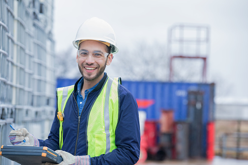 Young adult Hispanic man is chemical engineer, checking inventory of chemical shipment at oil and gas industry job site. Pipeline pipes and other equipment are in background. Man is wearing safety gear and using a clipboard.