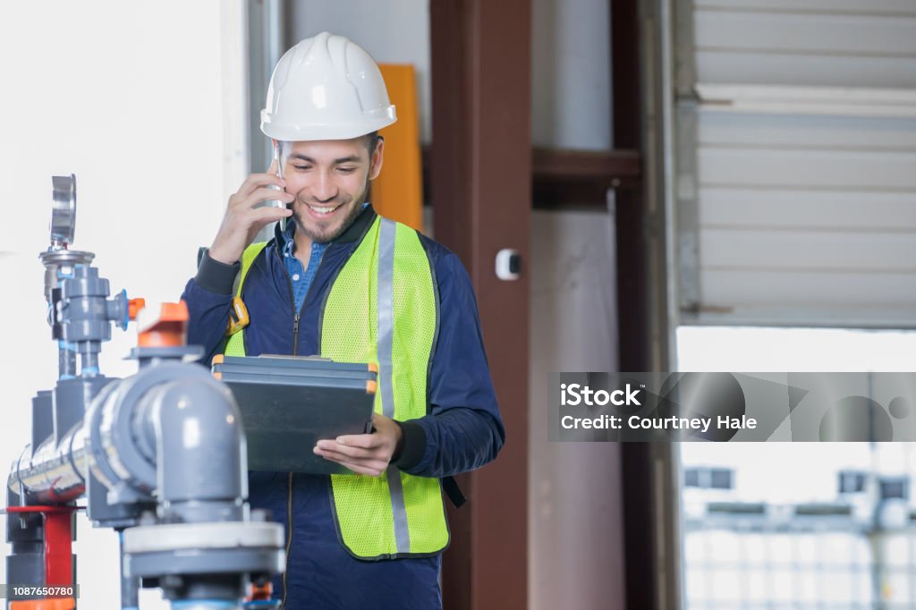 Engineer working in oil and gas industry inspecting pipeline equipment on job site Young adult Hispanic man is engineer working in oil and gas industry on pipeline job site. Man is making phone call while inspecting equipment. Oil Pump Stock Photo