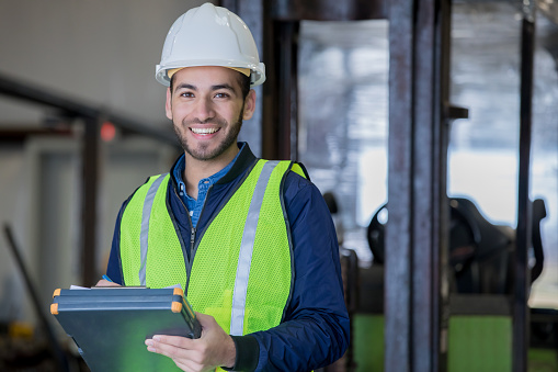 Young adult Hispanic man is smiling and looking at the camera while working in warehouse as forklift operator. Man is using a clipboard and wearing a hard hat and safety gear while standing in front of forklift.