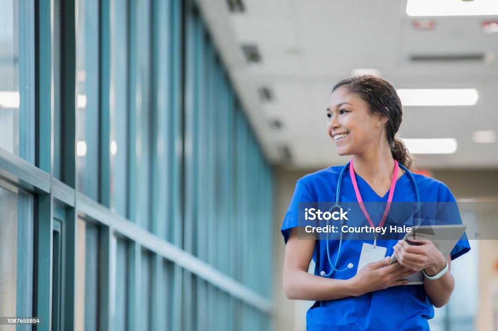 Happy young nurse smiles while walking in hospital sky walk during shift. Happy young adult mixed race woman is nurse or healthcare worker. She is carrying a digital tablet patient chart and looking out the window of sky walk in large hospital. Nurse Stock Photo