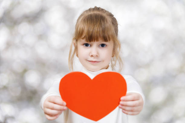 A small child is showing a heart in her hands. A tender smiling child is showing a big bright red heart in her hands at the light blurred background. The concept is the children's rights defending principle. childrens rights stock pictures, royalty-free photos & images