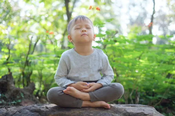 Photo of Cute little Asian 2 - 3 years old toddler baby boy child with eyes closed, barefoot practices yoga & meditating outdoors