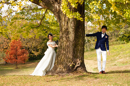 There is a tree standing at a park. An adult couple are standing by the tree. They wear formal wedding dress. They are just married.