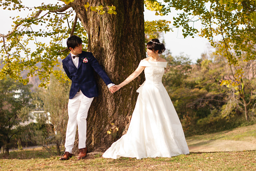 An adult wedding couple wares wedding dress and standing by a tree together. They are looking at each other and keeping hands there.