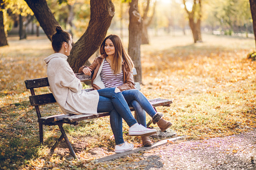 Two female friends talking on a bench in autumn park