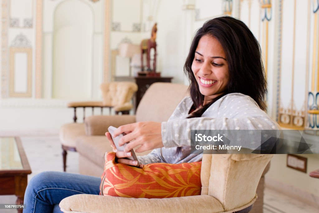 Young indian woman reading funny messages on mobile phone Happy smiling young indian woman sitting on sofa in hotel lobby having fun reading messages on her mobile phone. Jodhpur, India. Culture of India Stock Photo