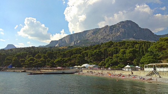 Picturesque view on Foros beach from a pier.
Sea in the foreground, some people on the beach.
Thick woodland stretched down to & along the beach on a background of a tremendous mountain ridge in a bluish haze..