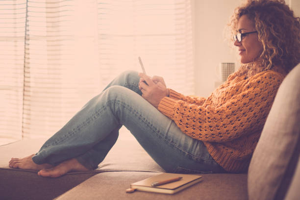 beautiful middle age caucasian woman at home lay on the sofa using mobile phone internet technology - paper note and pen on the sofa - barefoot enjoying the living room - vintage warm colors