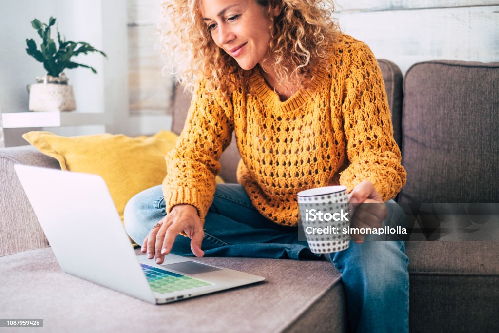 Nice beautiful lady with blonde curly hair work at the notebook sit down on the sofa at home - check on oline shops for cyber monday sales - technology woman concept for alternative office freelance Nice beautiful lady with blonde curly hair work at the notebook sit down on the sofa at home - check on line shops for cyber monday sales - technology woman concept for alternative office freelance Women Stock Photo