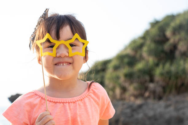 A young girl of mixed race poses with a pair of paper star-shaped glasses at the beach.