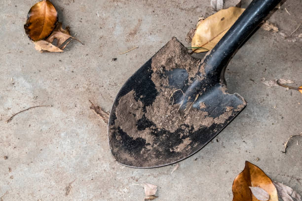 A black shovel with dirt caked on it from digging lays on a sandy concrete surface with brown winter leaves scattered around A black shovel with dirt caked on it from digging lays on a sandy concrete surface with brown winter leaves scattered around grave digger stock pictures, royalty-free photos & images