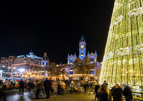 Valencia, Spain. December 2018: Christmas fair with carousel on Modernisme plaza of the city hall of Valencia at nightfall illuminated by christmas lights and a shinny christmas tree. Slow shutterspeed.