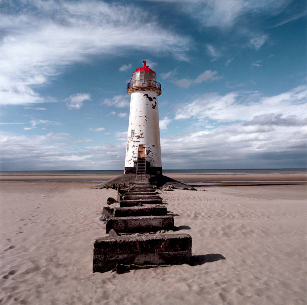 An old white light house with a bright red roof sitting on a sandy beach with a stone pathway infront, the sea is in the distance and a big blue sky with white swirling clouds is behind the light house, Talacre lighthouse, t