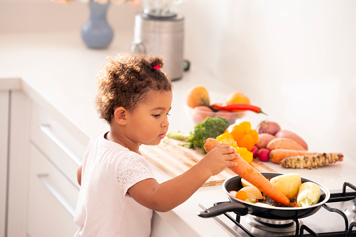 Little baby girl playing with vegetables, putting carrot and other roots on a pan, preparing food on the kitchen countertop.
