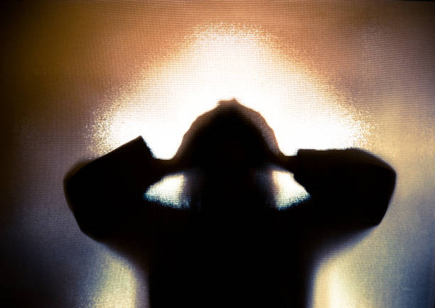 Silhouette of depressed abused woman with hands on head Colour backlit image of the silhouette of a woman with her hands on her head in a gesture of despair. The silhouette is distorted, and the arms elongated, giving an alien-like quality. The image is sinister and foreboding, with an element of horror. It is as if the 'woman' is trying to escape from behind the glass. Horizontal image with copy space. trapped stock pictures, royalty-free photos & images