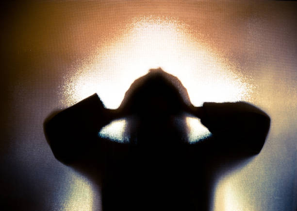 Silhouette of depressed abused woman with hands on head Colour backlit image of the silhouette of a woman with her hands on her head in a gesture of despair. The silhouette is distorted, and the arms elongated, giving an alien-like quality. The image is sinister and foreboding, with an element of horror. It is as if the 'woman' is trying to escape from behind the glass. Horizontal image with copy space. stuck stock pictures, royalty-free photos & images