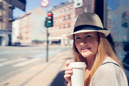 Female with long red hair wearing a hat having coffee to go  enjoying a walk in the city on a sunny day