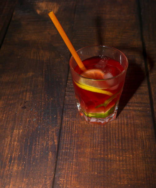 bicchiere umido di cocktail rosso freddo con succo di frutta, fette di lime, foglie di menta, cubetti di ghiaccio e paglia sulla scrivania di legno - studio shot gourmet freshness cold foto e immagini stock