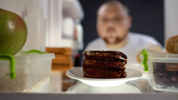 Oversize man taking piece of cake from fridge at night, diabetes risk, calories stock photo