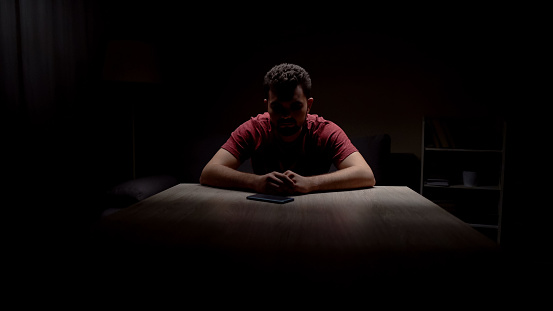 Deprimido solo hombre sentado en un cuarto oscuro mirando el teléfono, esperando llamada photo