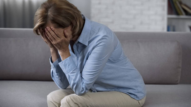 istock Aging lady sitting on sofa alone, covering face and crying, suffering headache 1088258816