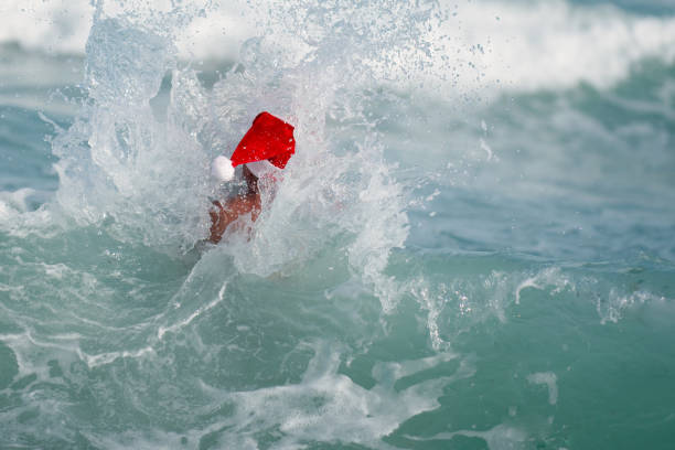 Senior caucasian ethnicity man is swimming in the water ocean. He is wearing a Christmas Santa Claus hat. The picture was taken in Florida USA. He is 60 years old. He is swimming in the waves. The water is blue and white.