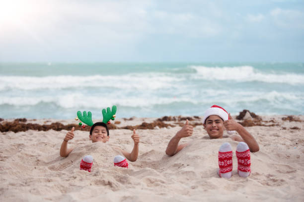 2 hispanic brothers having fun at the beach during Summer Christmas. They play in the sand together and wear some Christmas props such as Santa Claus hats and socks. Pictures were taken in Florida USA. One of them is generation Z. They are Mexican American and have brown hair.