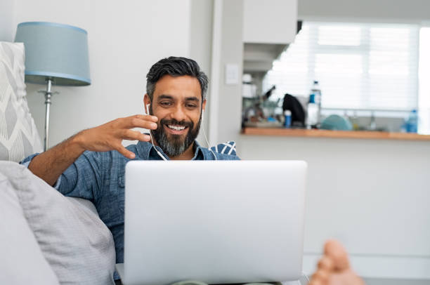 Man using laptop for video call Happy mature man relaxing on couch while video calling using laptop at home. Latin man sitting on sofa and making a video call. Smiling middle eastern businessman doing online video chat while gesturing with hands. asian man headphones stock pictures, royalty-free photos & images