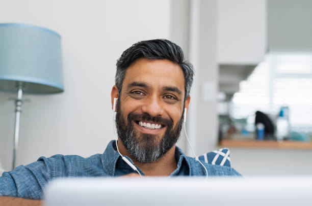 Happy man at home Portrait of happy man using laptop with earphones while lying on couch. Casual man in video call lying on sofa while listening to music with computer. Mature middle eastern man with earbuds looking at camera with a big smile. asian man headphones stock pictures, royalty-free photos & images