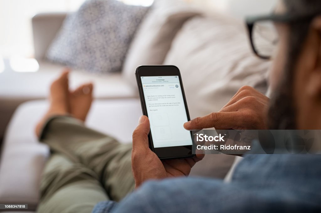 Man reading email on smartphone Closeup of a man hand holding cellphone with internet browser on screen. Man with spectacles relaxing sitting on couch while looking at mobile phone. Closeup of mature latin man using smartphone to checking email at home. Telephone Stock Photo