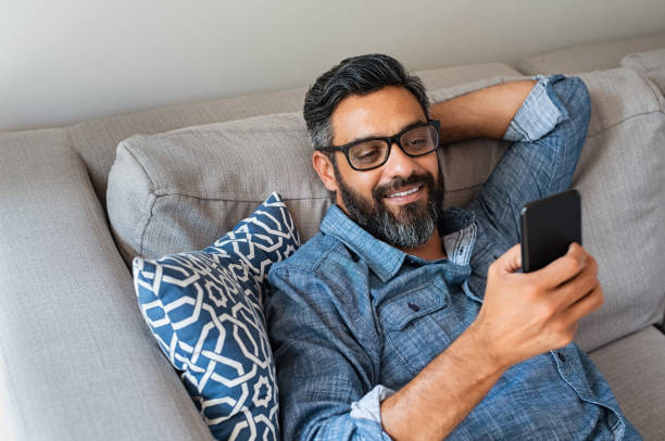 Man using smartphone at home Happy smiling latin man using smartphone device while sitting on sofa at home. Mature indian man lying on couch reading messages on mobile phone. Hispanic guy with eyeglasses looking at cellphone while relaxing at home. smirking stock pictures, royalty-free photos & images