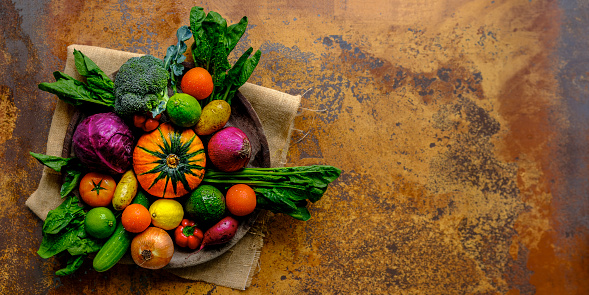 Many colorful contrast color salad vegetables and lemon and lime, sitting in a round, old, wooden vegetable bowl on burlap sack material on an abstract terracotta rustic background with atmospheric lighting, with good copy space to the right of the image.