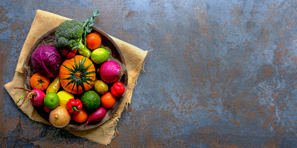 Many colorful contrast color salad vegetables and lemon and lime, sitting in a round, old, wooden vegetable bowl on burlap sack material on an abstract blue/grey rustic background with atmospheric lighting, with good copy space to the right of the image.