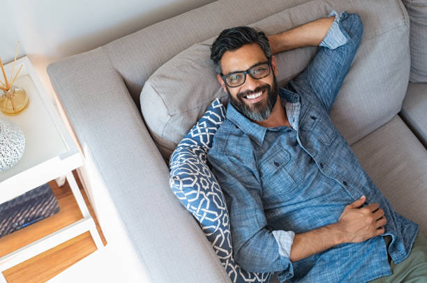 homme relaxant souriant à la maison - hands behind head photos et images de collection