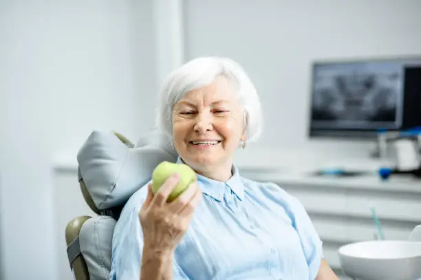 Portrait of a beautiful senior woman with healthy smile holding green apple at the dental office