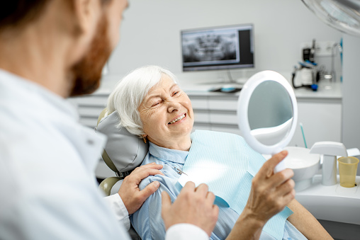 Happy elderly woman enjoying her beautiful toothy smile looking to the mirror in the dental office