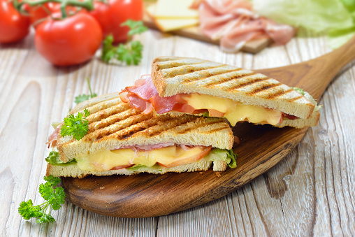 Grilled and pressed toast with smoked ham, cheese, tomato and lettuce served on sandwich paper on a wooden table