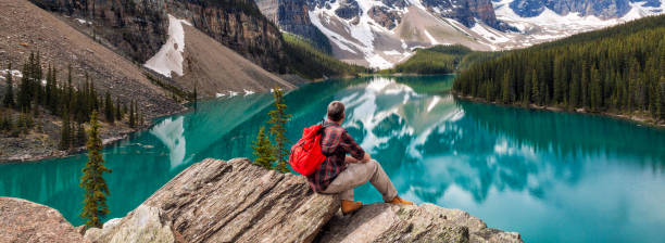 istock Panoramic web banner hiking man sitting down with rucksack backpack standing on tree log by Moraine Lake looking at snow covered Rocky Mountain peaks, Banff National Park, Alberta Canada 1088417386