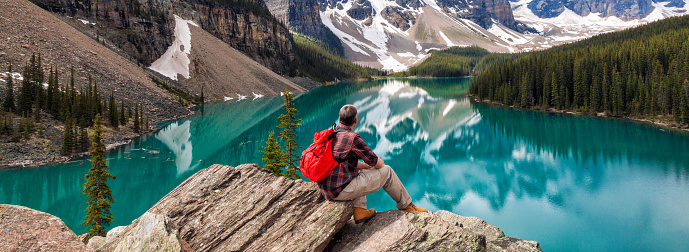 Panoramic web banner hiking man sitting down with rucksack backpack standing on tree log by Moraine Lake looking at snow covered Rocky Mountain peaks, Banff National Park, Alberta Canada