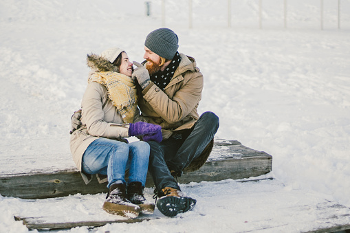 Loving couple young people a man with a beard and a Caucasian woman are sitting on a wooden staircase in the winter on the snow St. Valentine's Day a date Christmas hugging and happy.