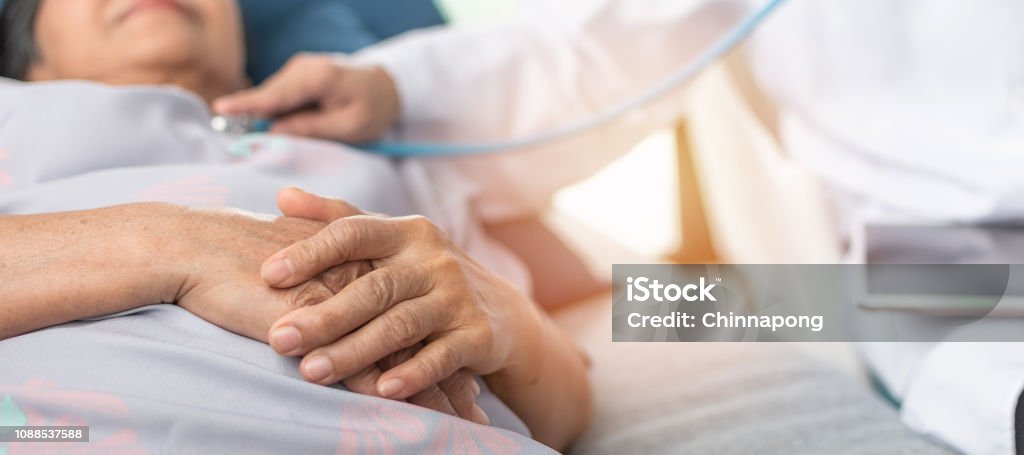Hospitalized elderly patient, senior old aging woman laying on bed with cardiologist doctor or physician examining cardiological heart health, checking pulse in hospital clinic exam room Hospital Stock Photo
